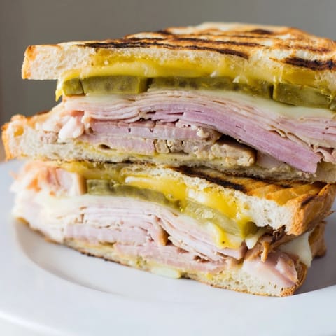 A toasted Cuban sandwich sliced diagonally, displaying warm layers of pork, ham, Swiss, and mustard on a cutting board.