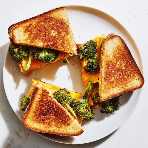 A comforting Roasted Broccoli & Cheddar Grilled Cheese rests beside a bowl of tomato soup, with fresh broccoli florets scattered on the rustic wooden table.